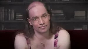 The image features a man sitting on a couch, wearing a pink floral top and glasses. He has long hair that falls over his shoulders. The background of the image shows a bookshelf filled with numerous books, suggesting an intellectual or academic setting. The caption at the bottom of the image reads "stop with this mumbo jumbo its not a real project $TIBBIR". This phrase is likely a reference to the man's appearance and possibly his involvement in some sort of fake or staged project.