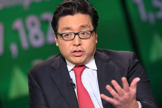 A man with glasses wearing a dark suit and red tie gestures with his hand while speaking, set against a green background with faint numerical text.