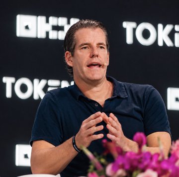 A man speaks into a headset microphone at the TOKEN2049 conference, with the "OKX" and "TOKEN" logos visible in the backdrop behind him. The image aligns with the post by showing one of the Winklevoss twins participating in a panel discussion about $ZEC at the event.