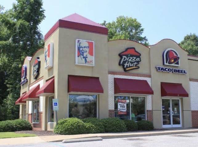 An exterior view of a strip mall featuring KFC, Pizza Hut, and Taco Bell restaurants, all sharing the same building with distinctive signage and red awnings.