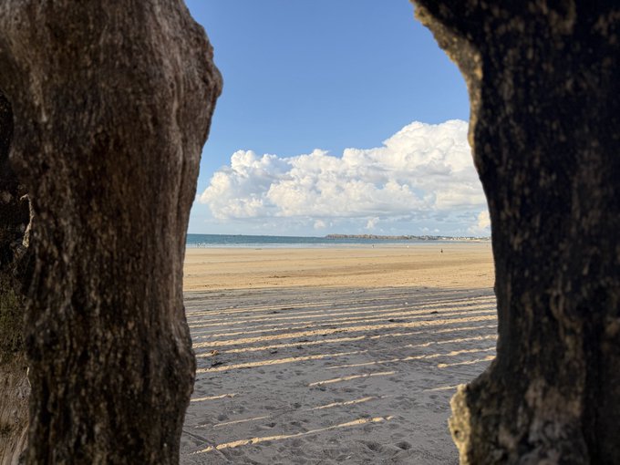 View of a sandy beach and ocean framed between two large rocks, with dramatic white clouds in a blue sky.