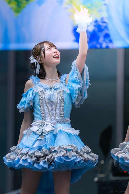 A young woman in an elaborate blue and silver frilled dress performs on stage, waving with one hand raised while smiling.