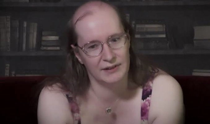 A balding person with glasses and long hair, wearing a floral top and a pendant necklace, sits in front of dark bookshelves in a dimly lit room.