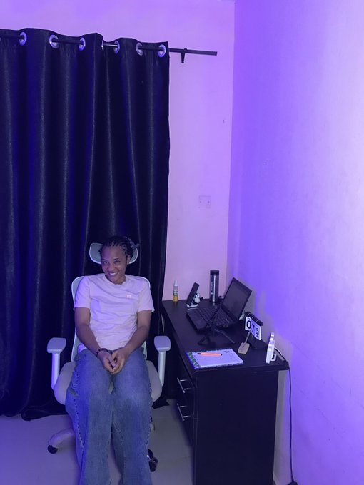A young woman sits smiling in a white office chair in a room with purple ambient lighting, next to a desk holding a computer monitor and electronics.