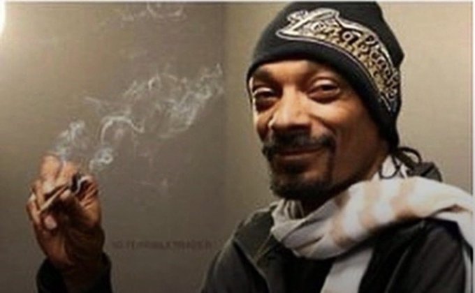A man wearing a black Los Angeles knit beanie and scarf smiles while holding a smoking cigarette.