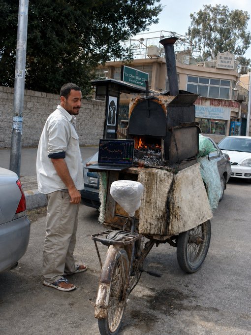 A man stands beside an improvised bicycle-mounted oven with a tall chimney, preparing to bake on a street in Egypt near signs reading "El Shamy Restaurant" and "City Center." The image of a street vendor hustling between jobs ironically pairs with the post's caption about checking Binance accounts between work, highlighting everyday hustle culture.