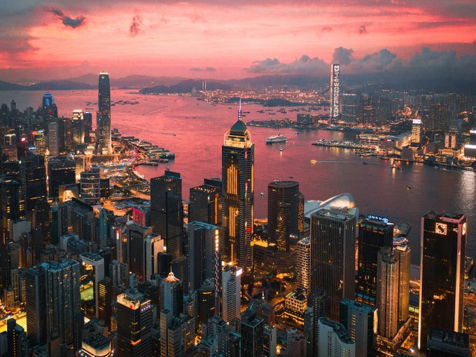 A dramatic aerial view of Hong Kong's skyline at dusk, with illuminated skyscrapers, Victoria Harbour, and a pinkish-red sky.