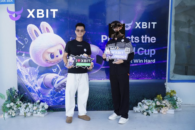 Two men pose with "I ❤️ XBIT" and "XBIT PREDICT" signs at an XBIT-sponsored event featuring a World Cup-themed backdrop with a pink bunny mascot and floral arrangements, with the XBIT logo prominently displayed. The post thanks gigiz_eth for the invitation and XBITDEX for sponsoring the event.