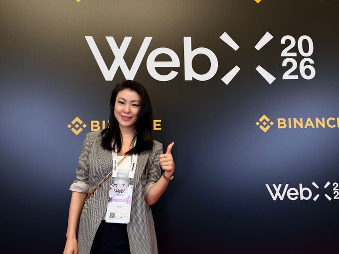 A woman wearing a lanyard and a plaid blazer smiles and gives a thumbs-up in front of a black backdrop featuring the white 'WebX 2026' logo and the yellow 'BINANCE' logo.