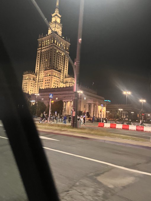 A nighttime view from inside a moving vehicle of the illuminated Palace of Culture and Science in Warsaw, Poland, with pedestrians on the sidewalk and a red-and-white construction barrier visible.