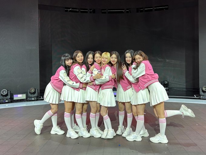 A group photo of nine young women in matching pink and white varsity jackets, pleated skirts, and white knee-high socks, posing together on what appears to be a stage.