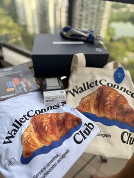 Two WalletConnect Club croissants-in-a-bag promotional items alongside a black box and HP laptop box on a table, with high-rise buildings visible through a window.