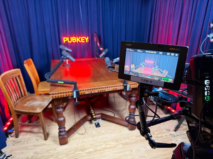 A podcast studio setup featuring a wooden table, microphones, blue curtains, a 'PUBKEY' neon sign, and a camera monitor labeled 'CAM A' displaying the recording in progress.