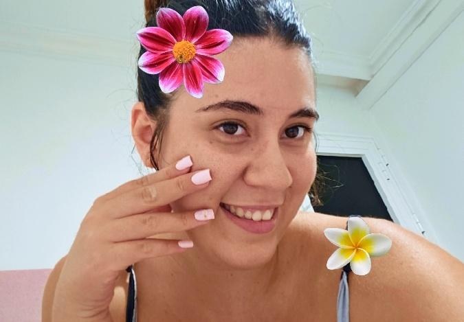 A smiling young woman with dark hair in a bun takes a selfie, digitally decorated with a pink flower filter on her head and a plumeria flower near her shoulder.