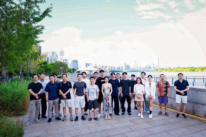 A group photo of approximately twenty casually dressed individuals standing on a paved waterfront promenade, with a city skyline and river visible in the background under a partly cloudy sky.