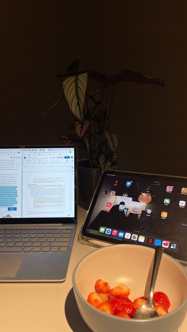 A dimly lit desk scene featuring an open laptop with a document on screen, a tablet displaying app icons, a potted plant, and a white bowl of strawberries with a spoon in the foreground.