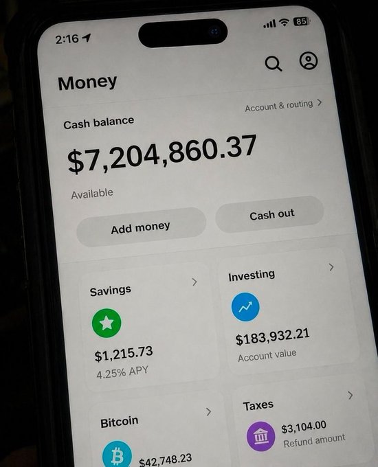 A photo of a smartphone screen displaying a banking app with a cash balance of $7,204,860.37, alongside savings of $1,215.73 at 4.25% APY, investing account value of $183,932.21, bitcoin worth $42,748.23, and tax refund amount of $3,104.00.