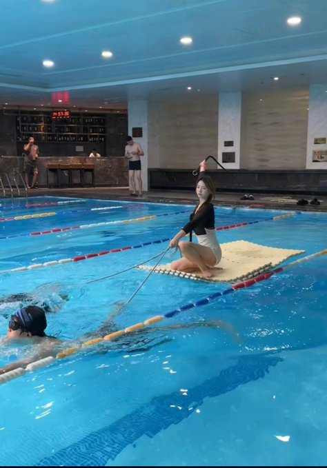 A woman in a black swimsuit sits on a floating platform in an indoor pool, holding a tether attached to a swimmer in the water, with other people visible in the background.
