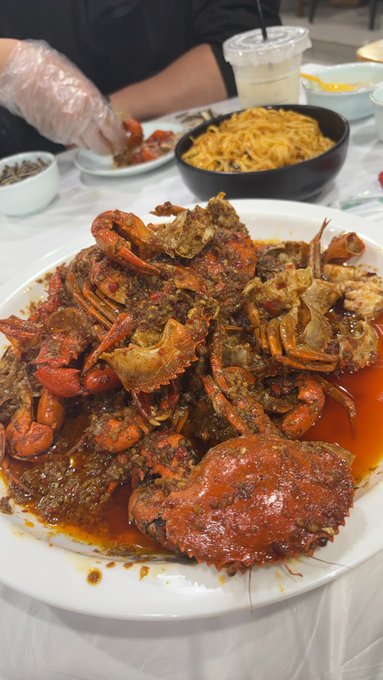 The image shows a plate of food with several crabs, including one that appears to be overcooked or burnt. The person sitting at the table has their hand covered by plastic gloves while eating the meal. There are also other plates of food on the table, indicating that this is likely a restaurant setting where multiple dishes are being served.