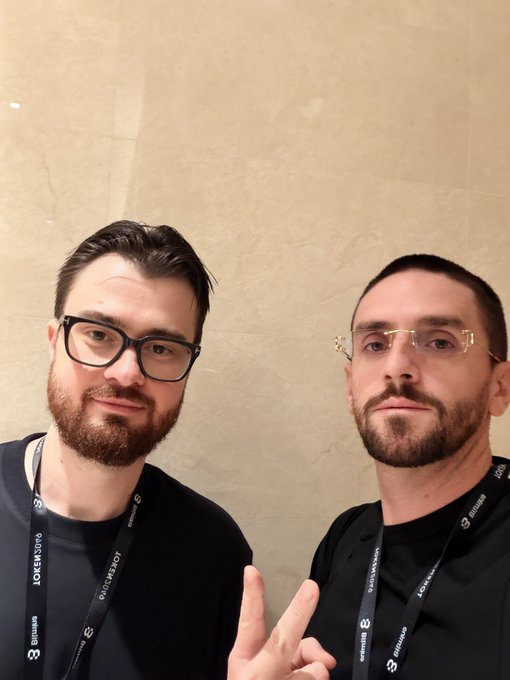 Two bearded men with glasses pose for a selfie, one flashing a peace sign, while wearing lanyards displaying "TOKEN2049" and "ODYSSEY.HOT" branding—tying into the post's reference to crypto figure Murad at a major crypto conference and the bullish $SPX6900 sentiment.