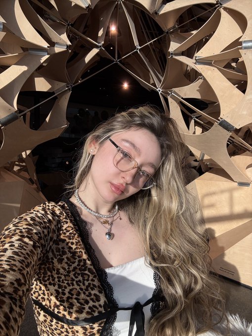 A woman with glasses and blonde hair takes a selfie while wearing a leopard print top and a heart pendant necklace, with a geometric wooden sculpture structure behind her.