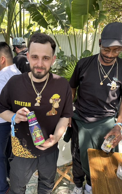 Two men pose at an outdoor gathering; the one in front wears a 'Cheetah' shirt with gold chains and holds a bottle, while the man behind him wears a black cap and shirt with chains.
