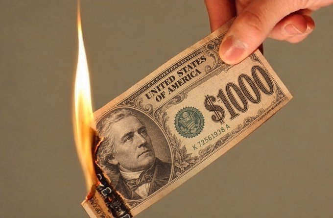 A hand holds a burning $1000 United States of America banknote, with the flame consuming the left edge featuring a portrait of a historical figure.