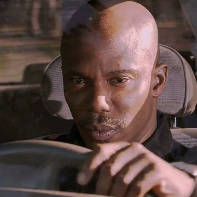 A close-up of a bald Black man with a serious expression sitting in a car, resting his hands together.