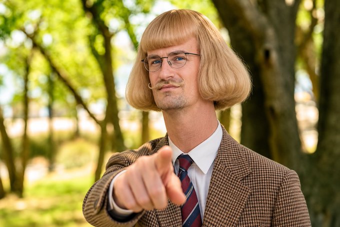 A man wearing a blonde bob wig, glasses, a mustache, a brown houndstooth blazer, white shirt, and striped tie, pointing directly at the camera outdoors.