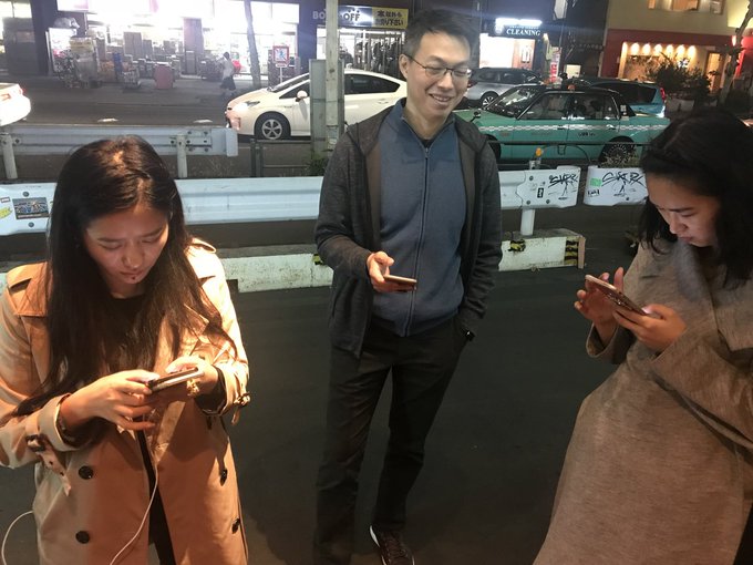 Three people stand on a Tokyo street at night, all focused on their smartphones, with taxis and storefronts visible in the background.