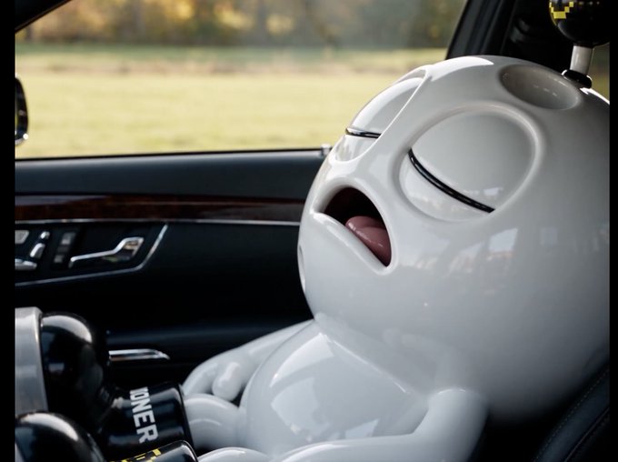 A glossy white cartoon-style figurine with closed eyes and an open mouth sits in the passenger seat of a luxury car, wearing a "JONES" seatbelt, with a countryside view visible through the window.