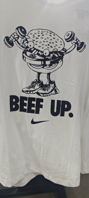 A white Nike t-shirt featuring a cartoon cheeseburger mascot lifting dumbbells with the bold text 'BEEF UP.' printed above the Nike swoosh logo.