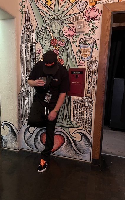 A person in a black t-shirt, shorts, and orange-accented sneakers poses in front of a colorful mural featuring the Statue of Liberty and city buildings.