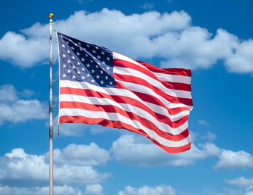 Photograph of the United States of America flag waving on a flagpole against a blue sky with scattered white clouds.