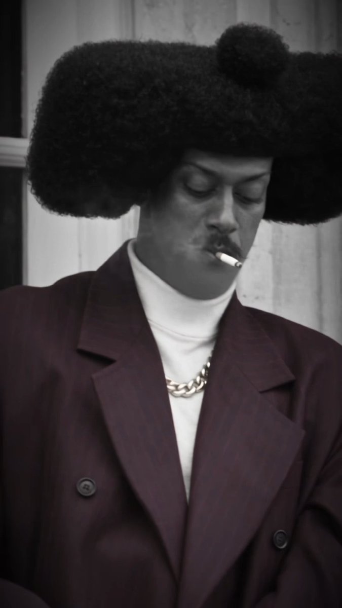 Black and white portrait of a person with a large, distinctive afro hairstyle, wearing a dark suit over a white turtleneck and a gold chain, with a lit cigarette hanging from their mouth.