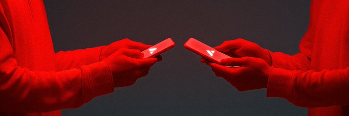 Two people in red hoodies face each other against a dark background, each holding a smartphone with an illuminated screen.