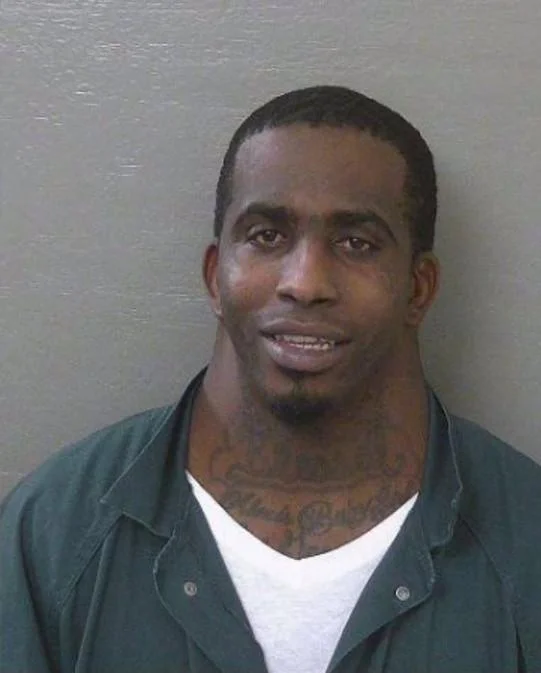 A mugshot of a Black man with short hair and a goatee, wearing a green prison uniform over a white shirt, with visible neck tattoos.