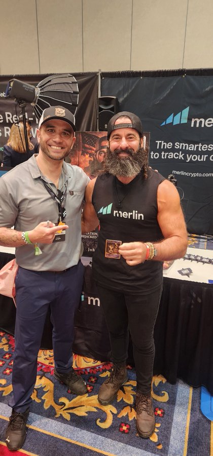 Two men pose together at XRP Las Vegas 2026 in front of a "merlin" (merlincrypto.com) booth, with the bearded man wearing a black "merlin" tank top and the other in a gray polo shirt, both smiling while standing on a colorful patterned carpet. The image captures a friendly networking moment from the cryptocurrency convention referenced in the caption, highlighting the community atmosphere Ripple fostered at the event.