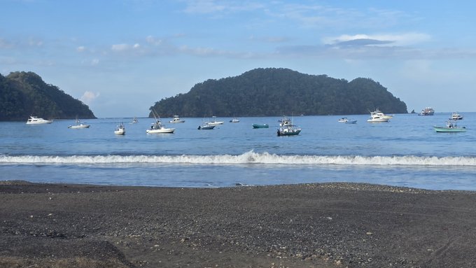 A scenic view of a dark sand beach in the foreground with small boats floating on the sea and forested hills/islands in the background under a partly cloudy sky.
