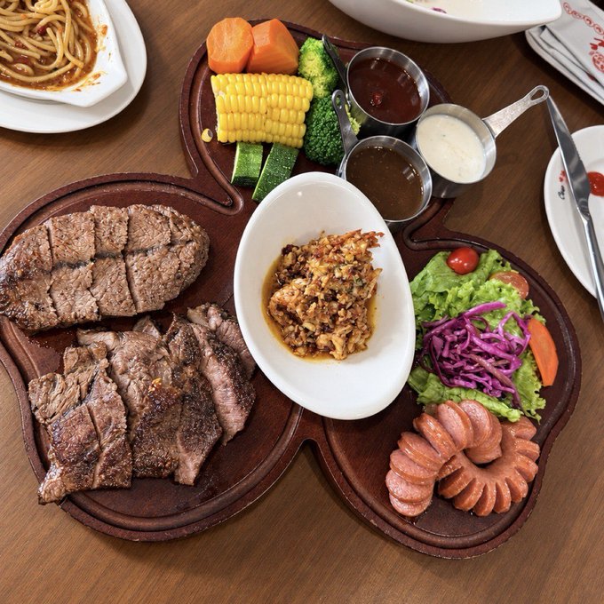 An overhead view of a steakhouse-style platter containing sliced steak, a hamburger steak, vegetables, corn, dipping sauces, salad, and sausage.