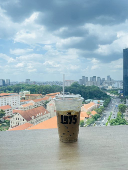 A plastic cup of iced coffee labeled "CAFÉ 1975" sits on a wooden ledge, overlooking a cityscape with traditional buildings and modern skyscrapers under a partly cloudy sky.
