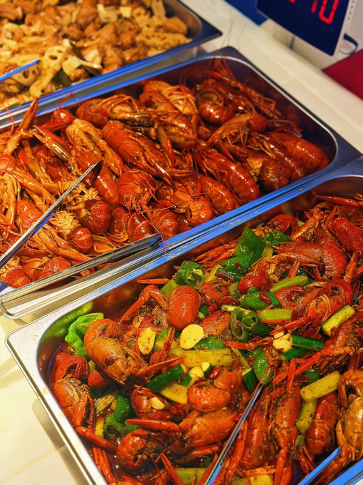 Close-up of a buffet spread of cooked red crawfish prepared in two different ways—one with green peppers and garlic, and another plain variety—with metal serving tongs resting in the trays.