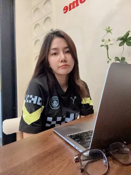 A young woman in an OKX-branded jersey sits at a desk with a laptop, looking unenthused about completing her monthly regulatory training—capturing the relatable struggle of balancing student life with workplace compliance requirements at the crypto exchange.