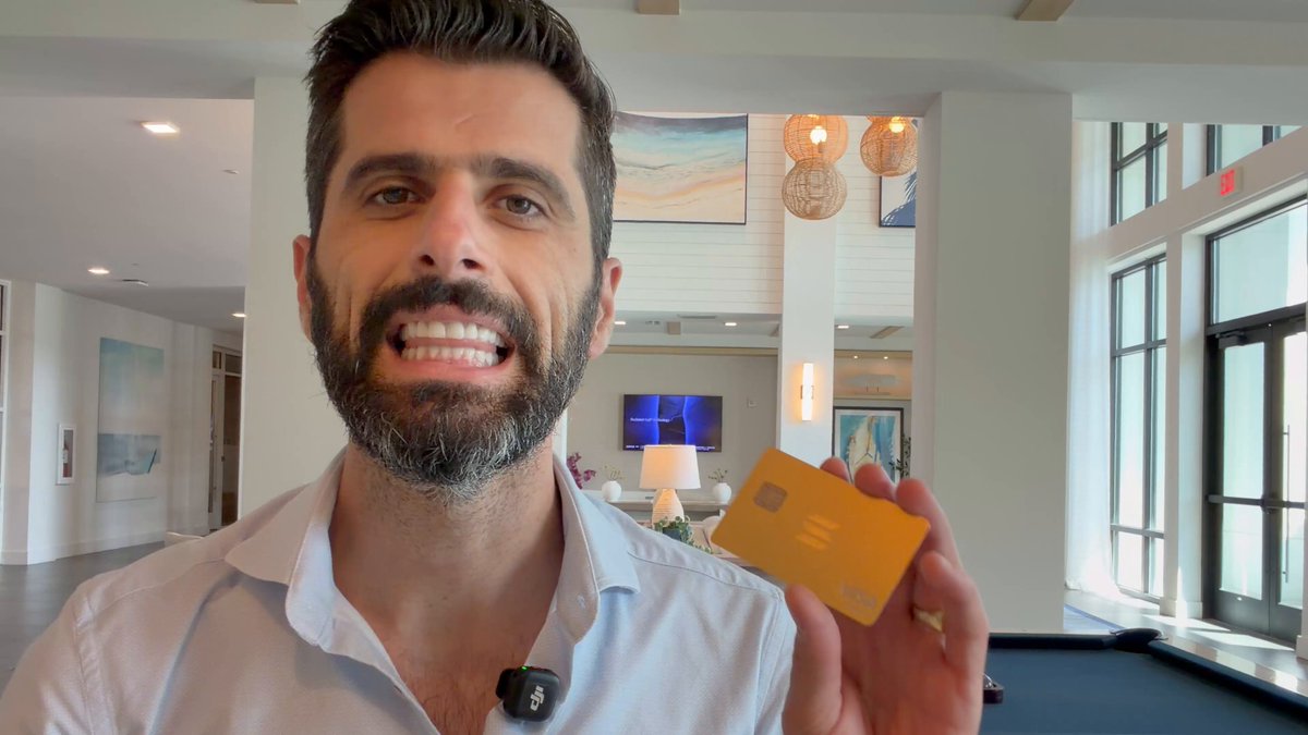 In this image, a man wearing a white shirt holds up an orange card, possibly a credit or debit card with a yellow stripe, suggesting he might be showing off some good spending or financial gain using cryptocurrency or a similar digital payment method. The photo is attributed to Kast Crypto Card creator Mr. John O'Connor. It appears that the image was taken by someone else and may not have been him directly but instead used for sharing his experience with the card.