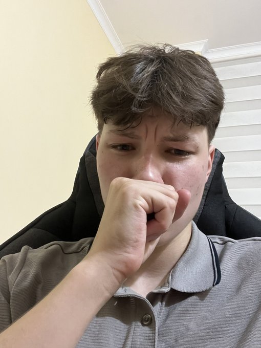 Close-up selfie of a young man with messy brown hair wearing a gray polo shirt, covering his mouth with his fist in an emotional expression, sitting in a black gaming chair with white window blinds in the background.