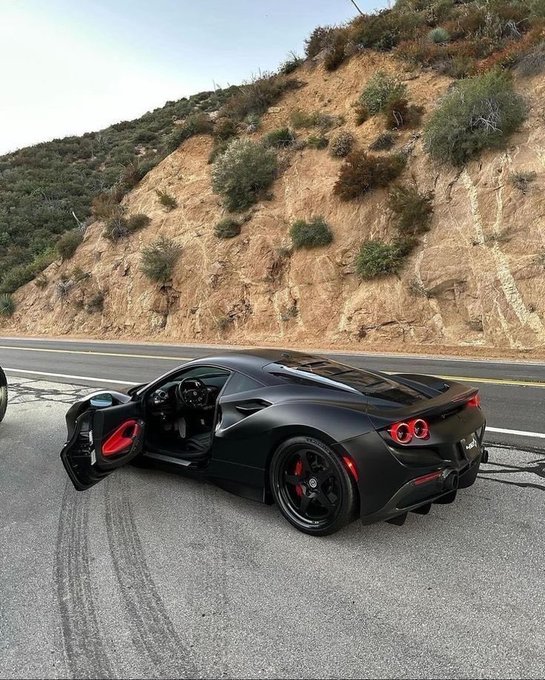A matte black sports car is parked on the side of a road with its driver's side door open, revealing a red interior, set against a backdrop of a dry, brush-covered hillside.
