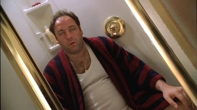 A man in a striped red and blue robe over a white tank top leans back in a doorway with gold trim, sporting a gold chain necklace. The caption "too many chains. too many coins." humorously references his flashy gold jewelry and robe, suggesting a gaudy display of wealth.