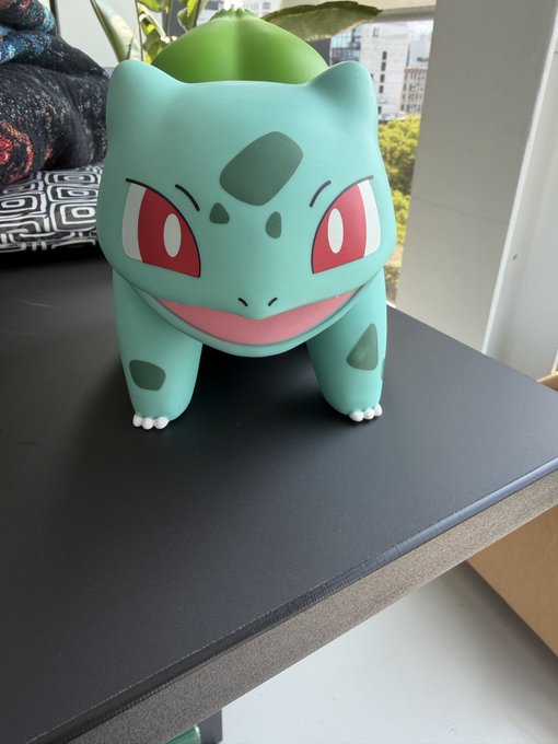 A 3D model or figurine of Bulbasaur, the green Pokémon with dark green spots and red eyes, sitting on a black desk near a window.