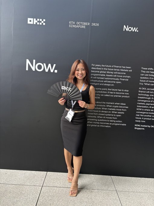 A woman in a black dress holding a branded fan stands in front of an OKX promotional backdrop dated "6TH OCTOBER 2026 SINGAPORE" with the word "Now." repeated.