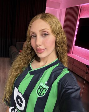 A young woman with curly blonde hair takes a selfie wearing a black and green striped polo shirt with "POLO" and a crest logo, appearing to lounge on a couch in a room lit with pink ambient lighting. The caption's mention of staying home aligns with the casual, indoor vibe, while the "make magic internet money" likely alludes to cryptocurrency trading or content creation.